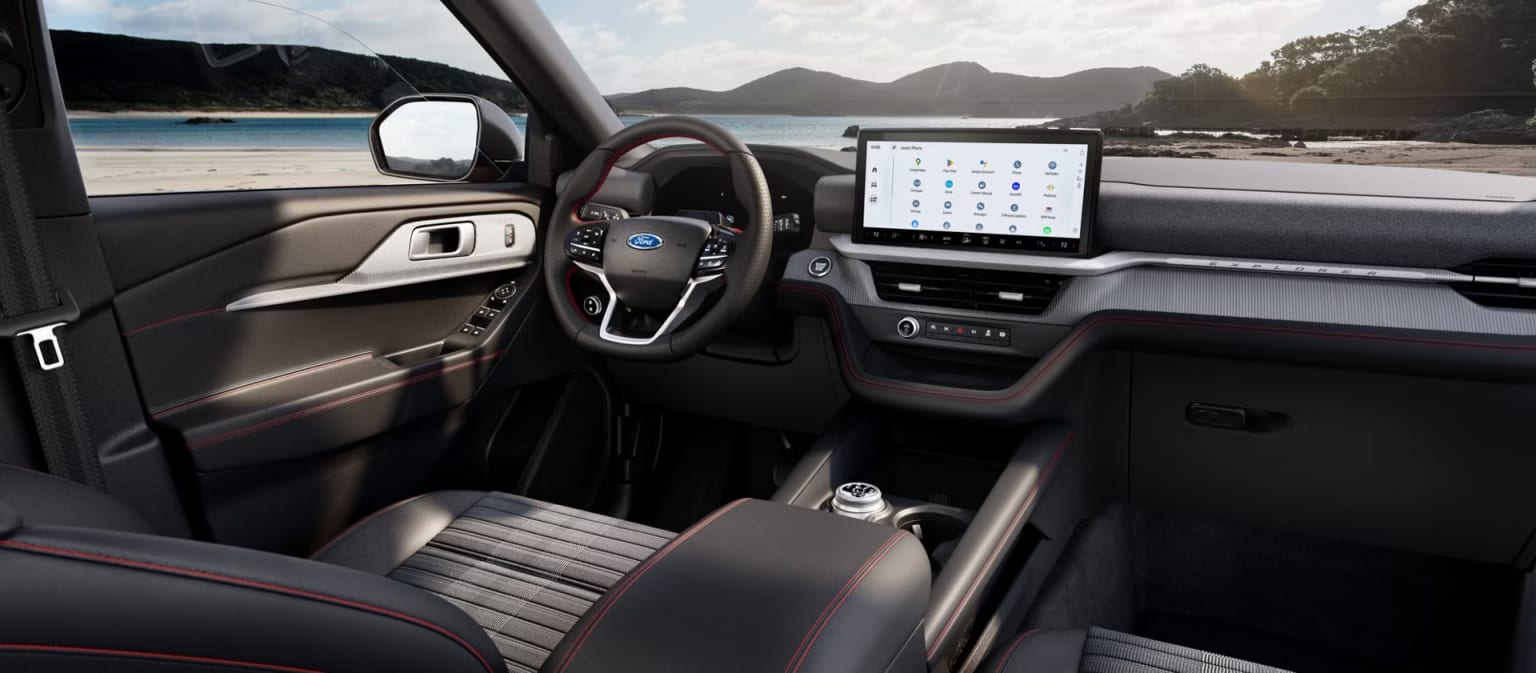 The interior of a modern vehicle, with a large touchscreen display and steering wheel, is visible in the foreground, while a scenic landscape with mountains and a body of water can be seen in the background through the windshield.