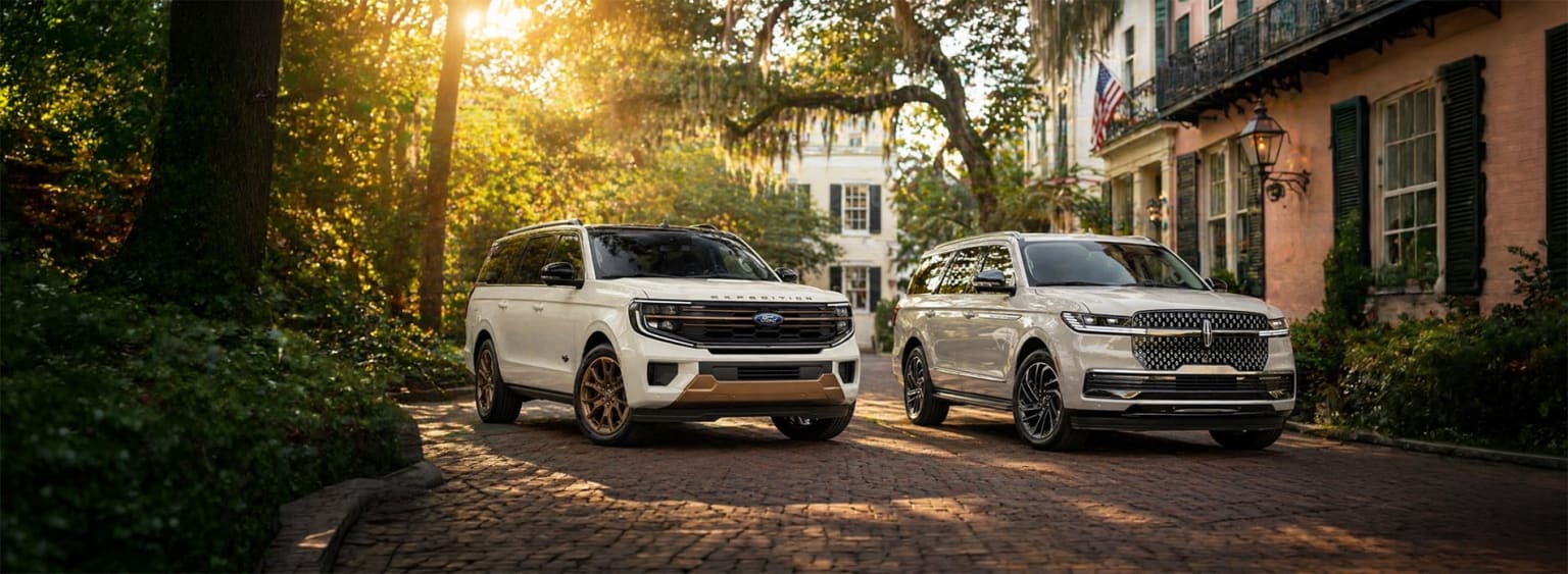 Two large, luxurious white SUVs parked on a cobblestone street, surrounded by lush greenery and sunlit buildings in the background.