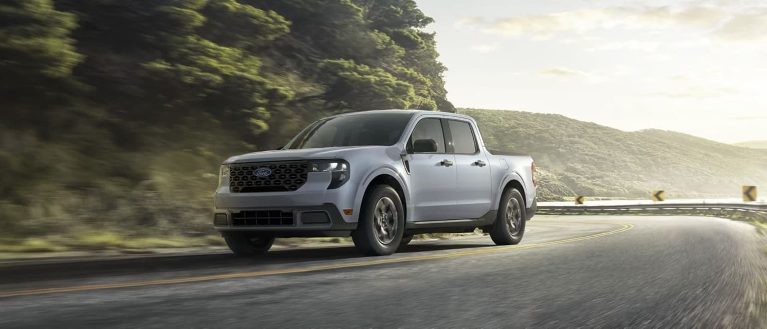 A pickup truck driving on a winding road surrounded by lush, green foliage and mountains in the background.