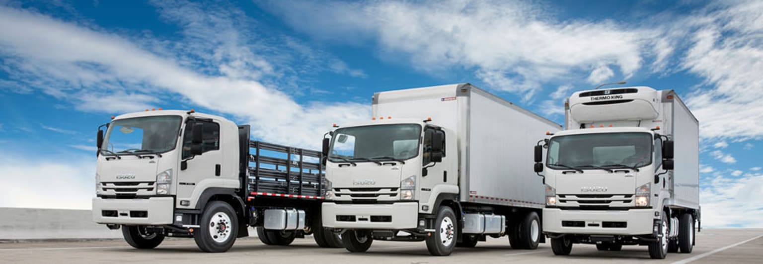 A fleet of commercial trucks, including a flatbed and box trucks, parked against a backdrop of a blue sky with fluffy white clouds.