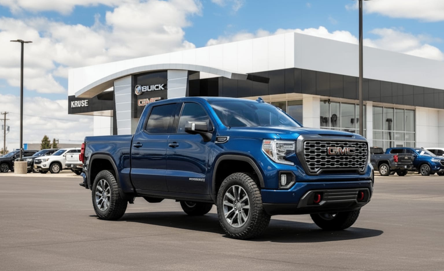 A large, rugged-looking blue pickup truck in foreground, modern, angular car dealership building in background against cloudy sky.