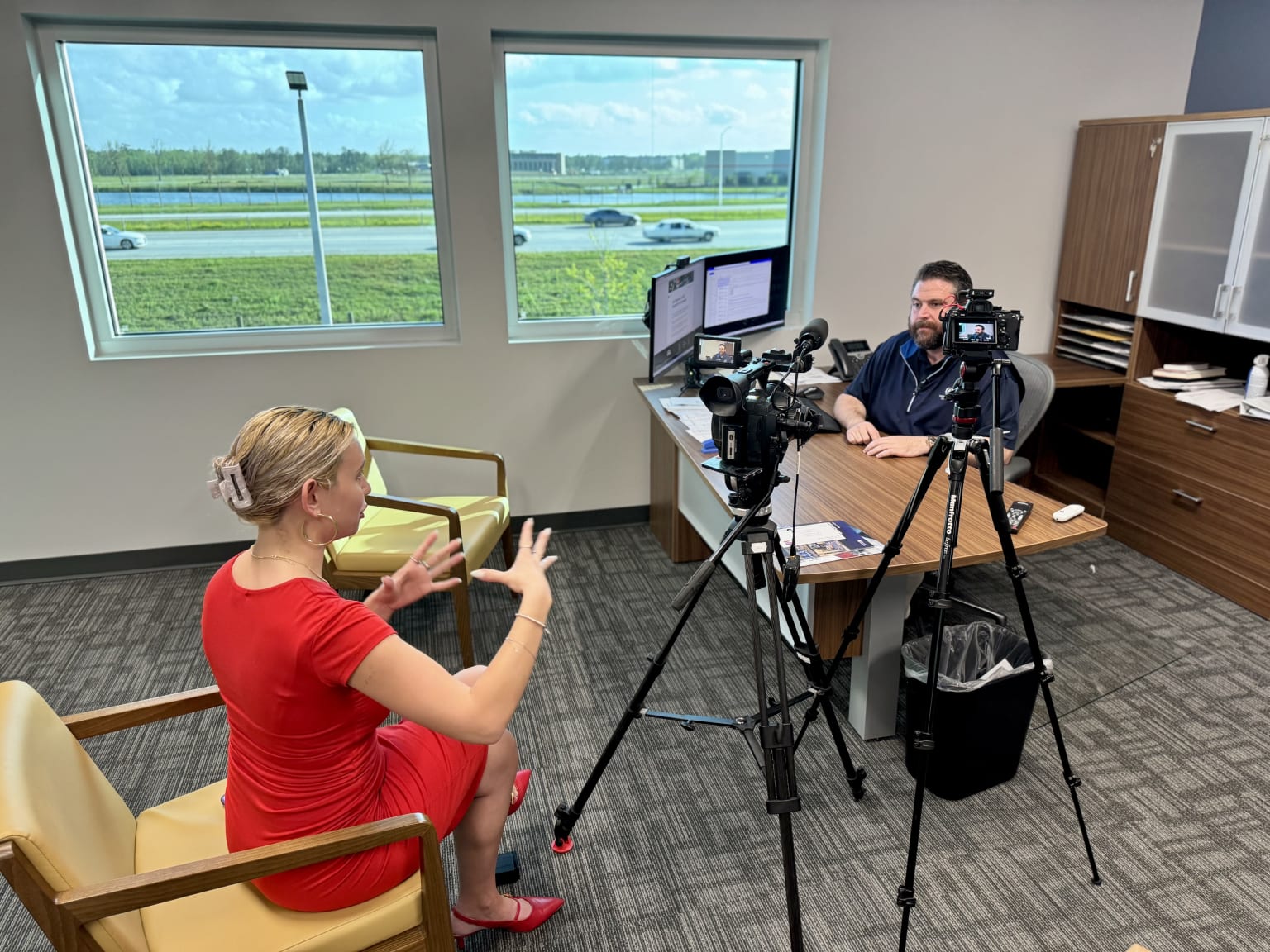 A woman in a red dress is seated in a chair, facing a camera operator who is filming her in an office setting with a scenic view of a grassy field visible through the windows.