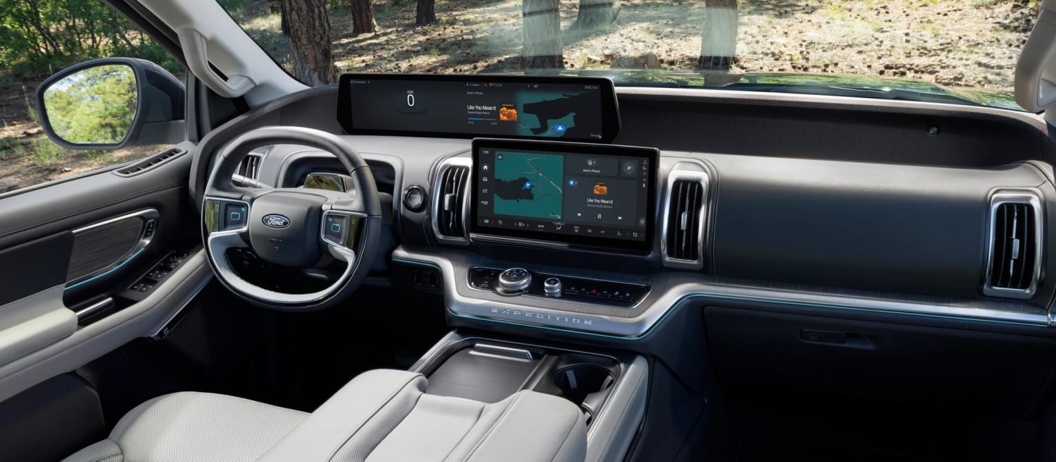 Interior of a modern vehicle with a sleek dashboard, multiple digital displays, and a steering wheel visible. Background outside the vehicle appears to be a forested area.