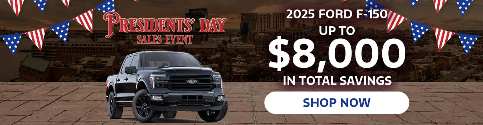 The image shows a large black pickup truck in the foreground, with a promotional advertisement for a Presidents' Day sales event in the background, featuring details about savings on a 2025 model.