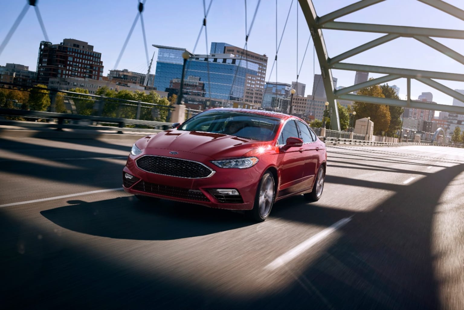 a red car driving on a bridge over a river with a cityscape in the background in the daytime