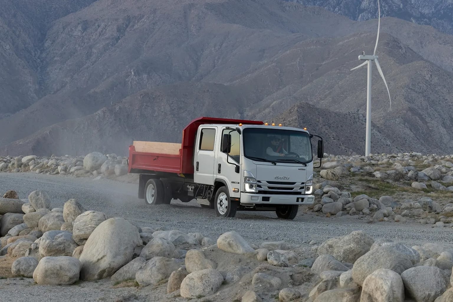 A large dump truck driving on a rocky mountain road with rugged terrain and a wind turbine in the background.