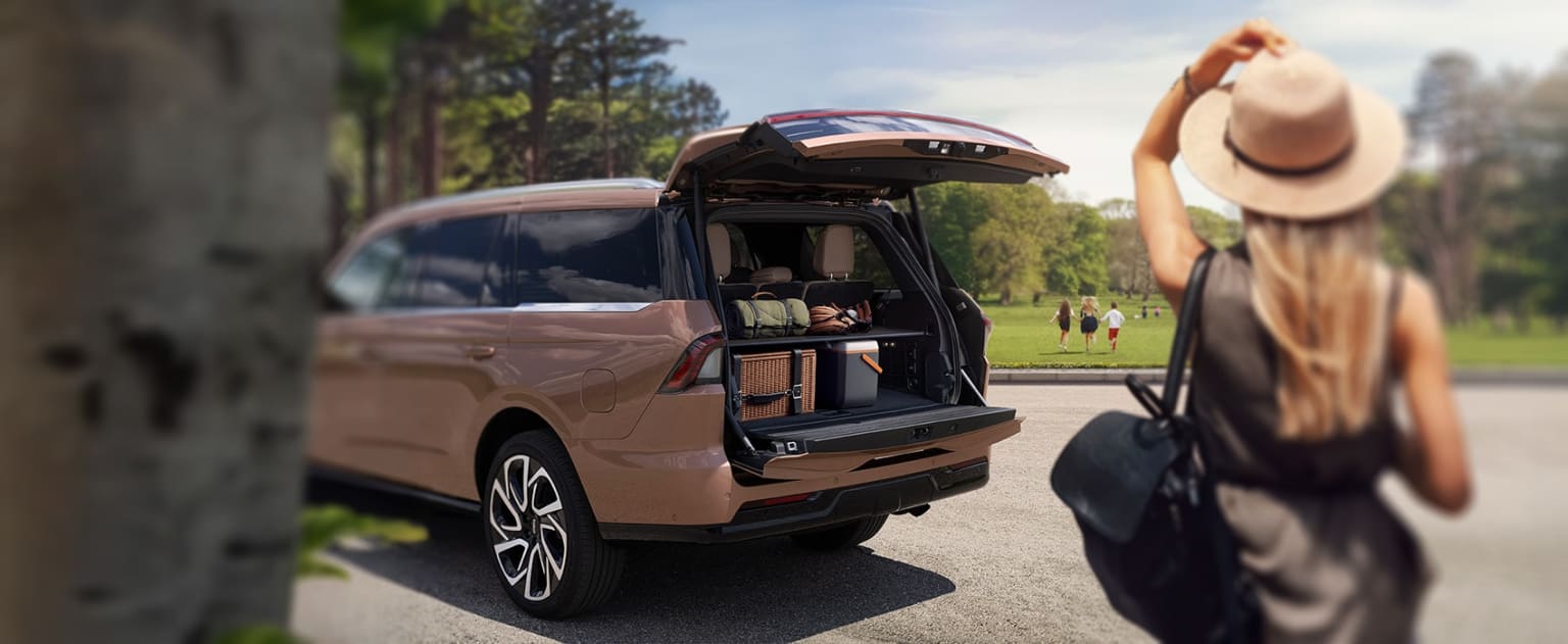 Beige SUV with an open trunk in a grassy area with trees in the background, person wearing a hat next to the vehicle.