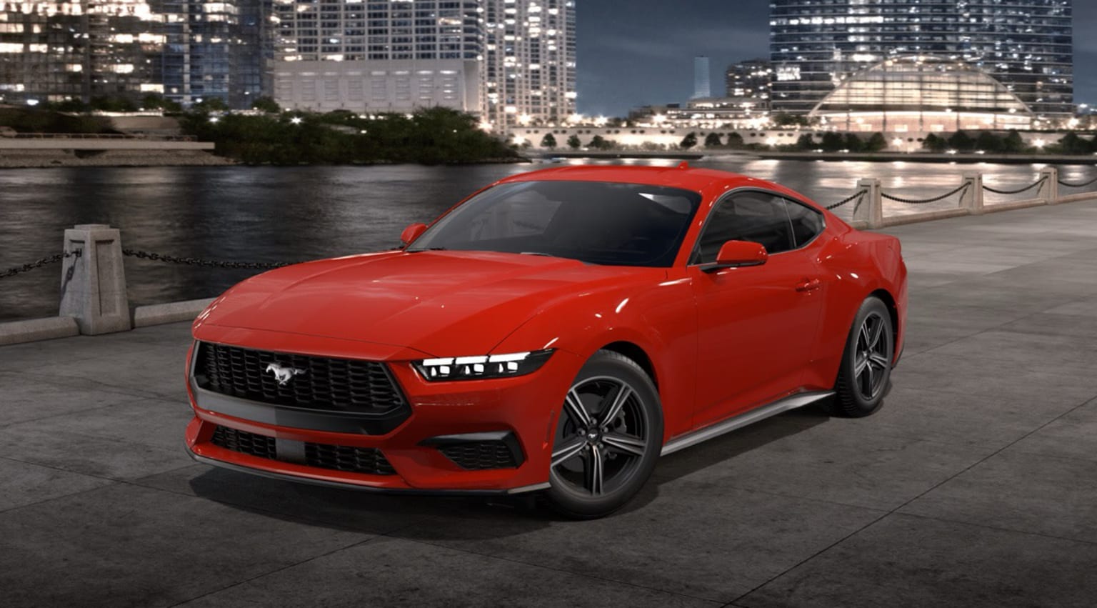 A red sports car parked on a waterfront promenade with a modern city skyline illuminated in the background.
