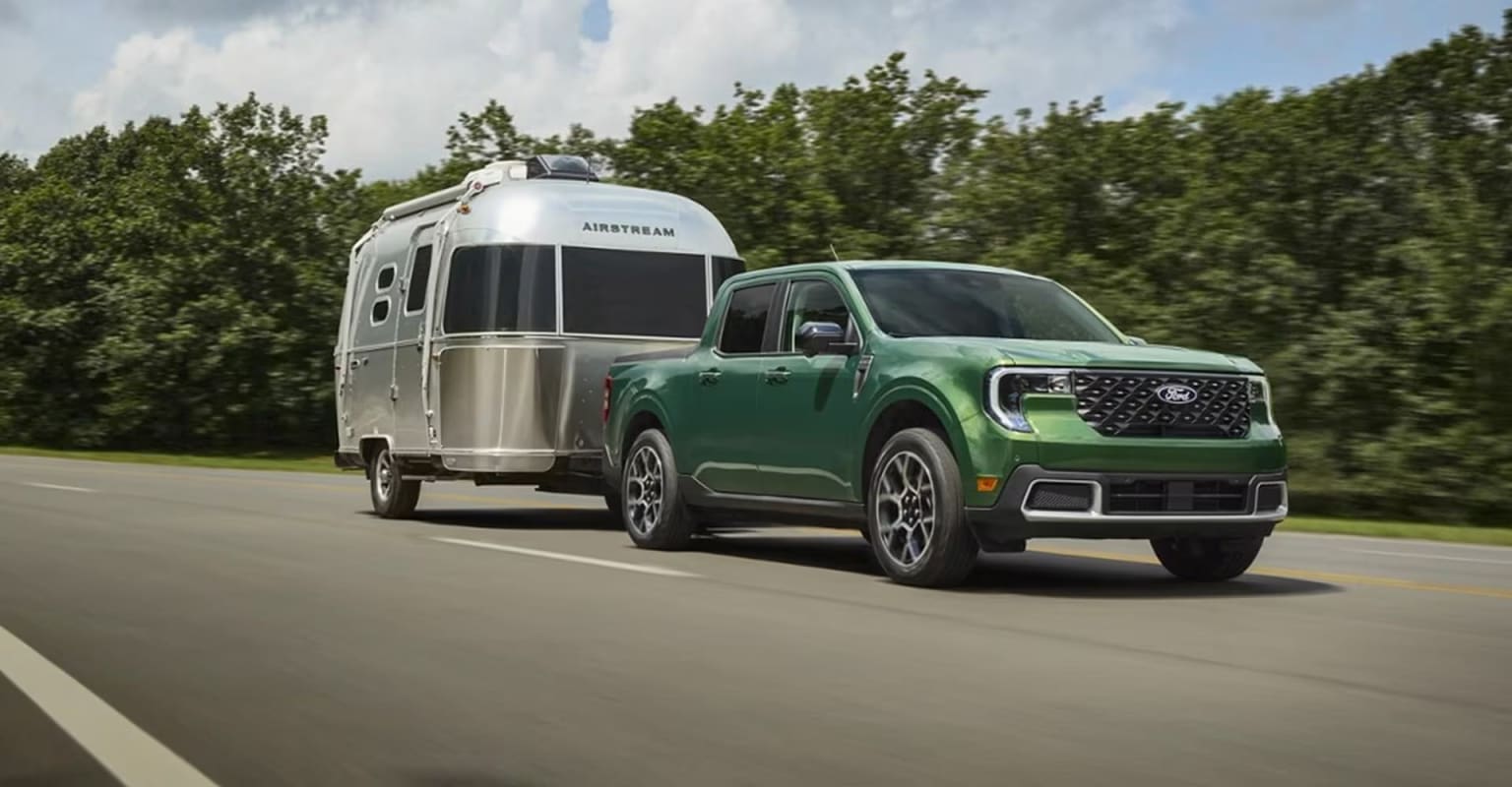 A green pickup truck is towing a silver Airstream trailer on a road surrounded by lush green trees.