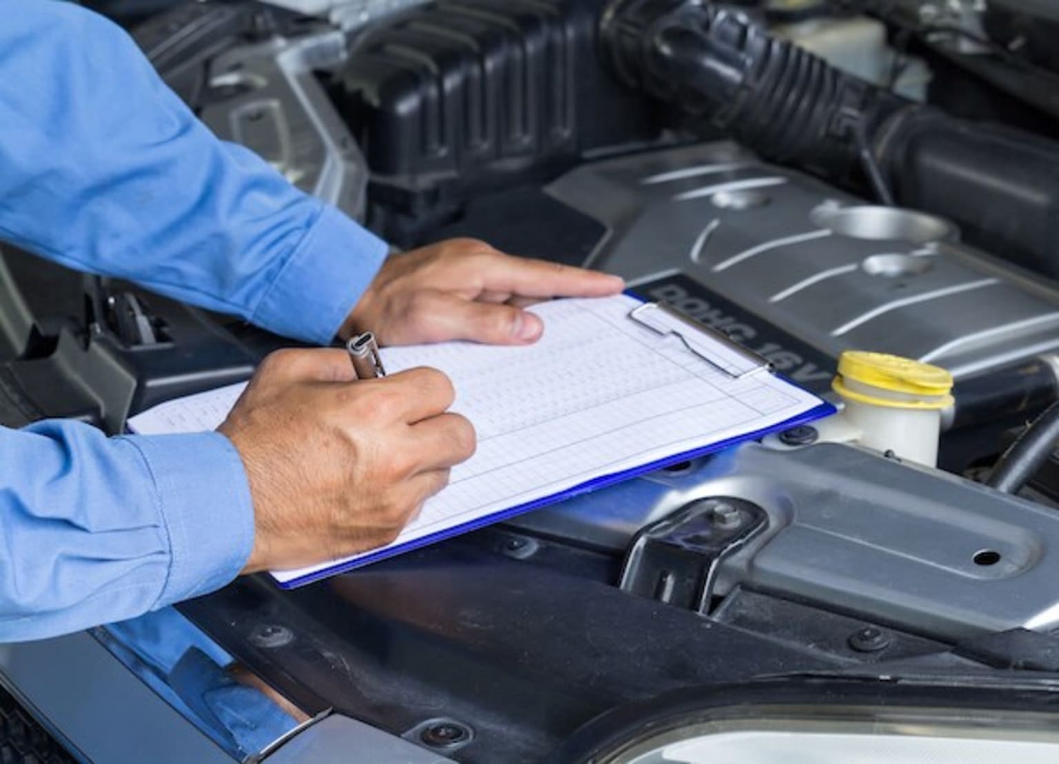 a man writing on a clipboard in front of a car's engine bay with the hood open