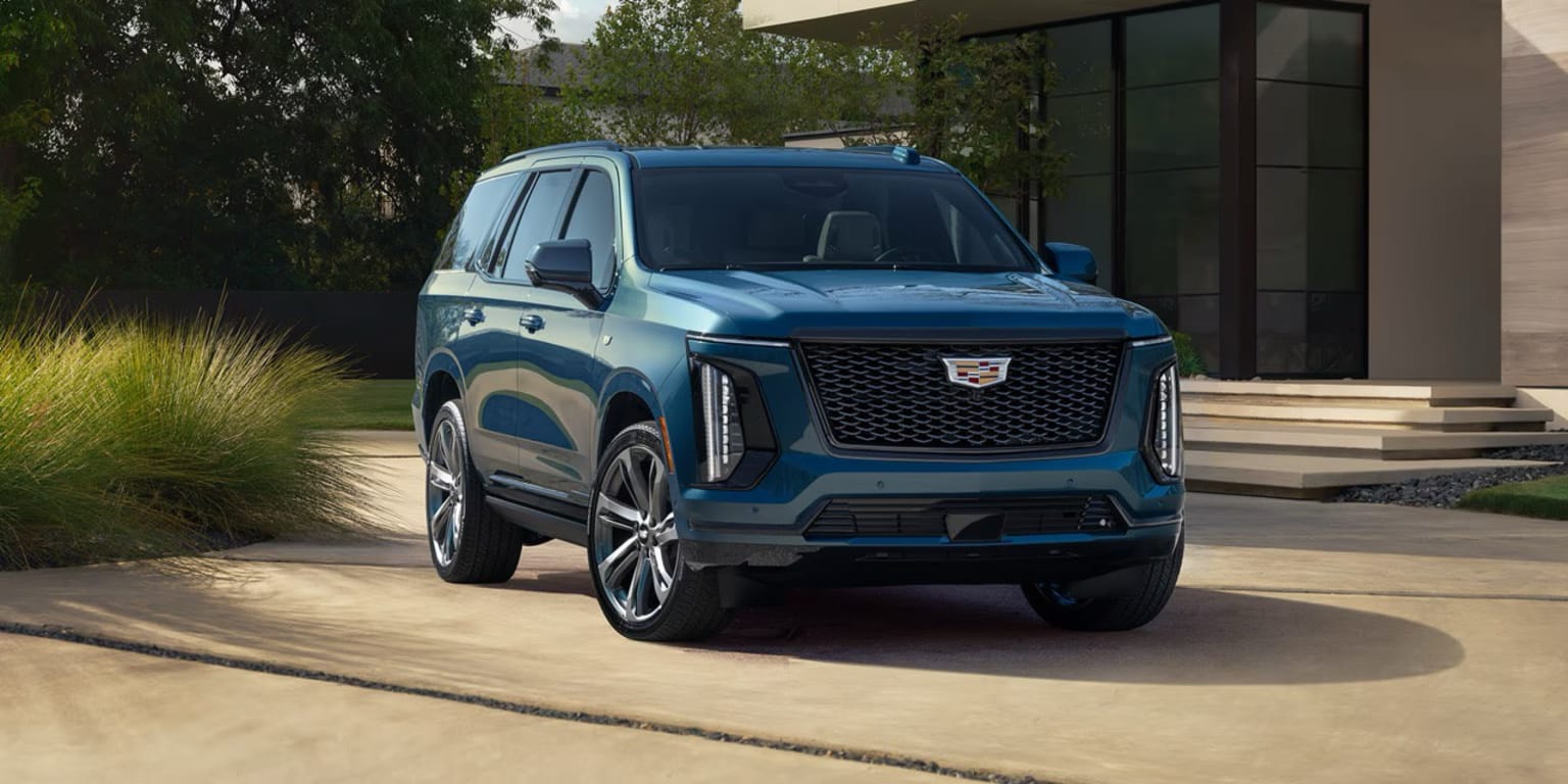 A large, blue SUV-style vehicle with a prominent grille and sleek design is parked in front of a modern building with a glass facade, surrounded by lush greenery.