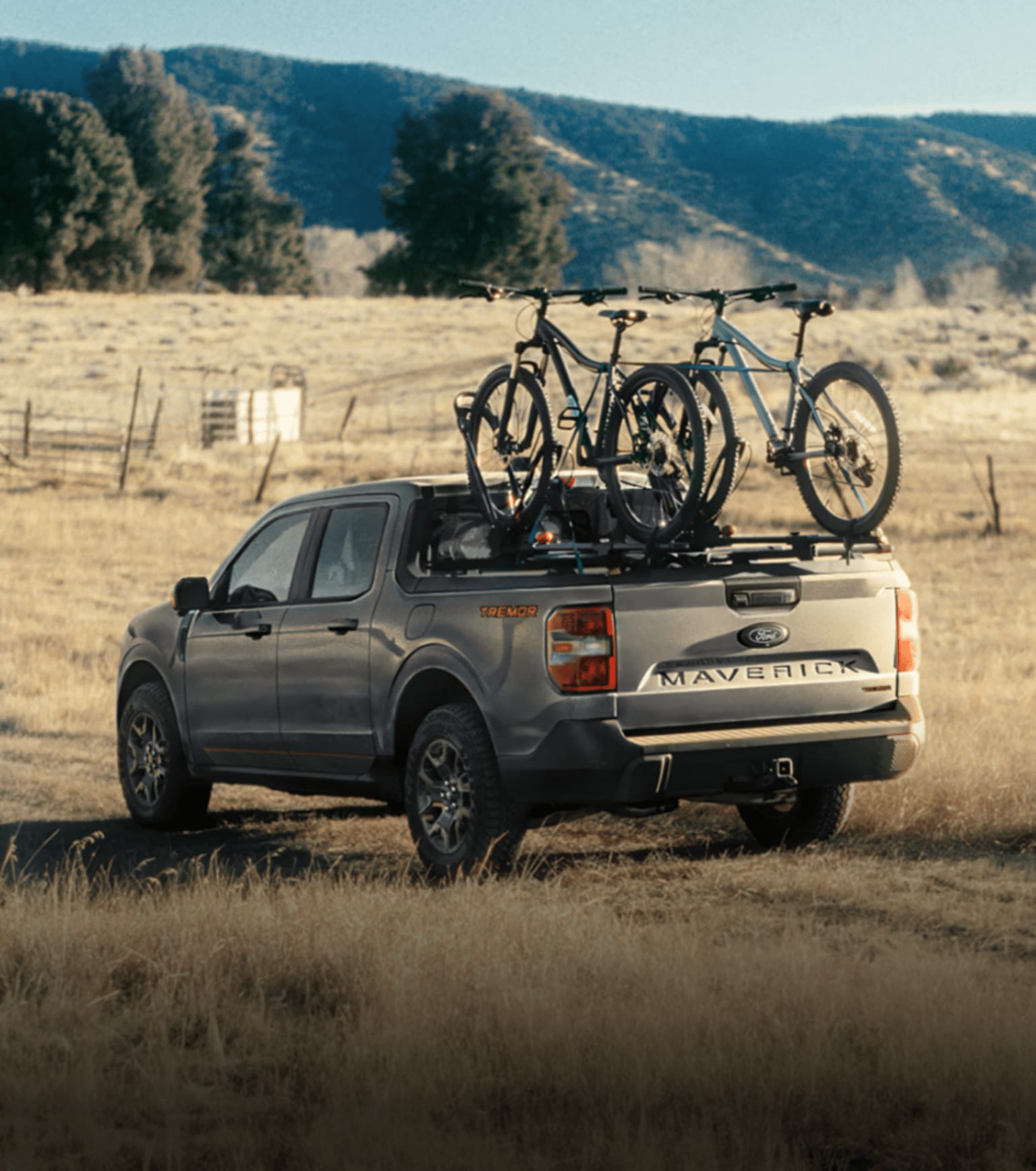 A grey truck with two mountain bikes in the bed parked in a grassy field with rolling hills in the background.