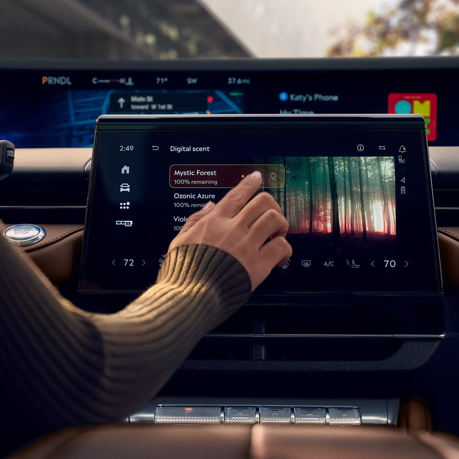 A person's hand interacting with the touchscreen display of a vehicle's dashboard, displaying various digital controls and information in the foreground, with a blurred outdoor scene in the background.