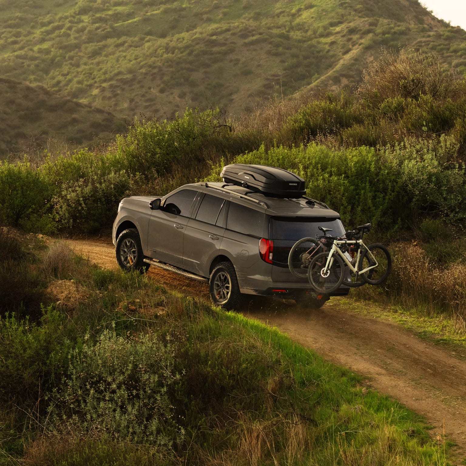 Off-road vehicle with roof rack and attached bicycle on a dirt path surrounded by lush, rolling hills covered in vegetation.
