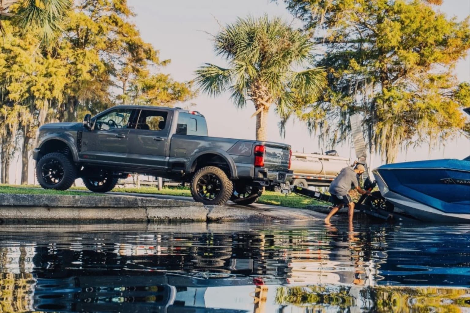 A dark gray pickup truck is parked on a concrete ramp next to a blue boat being pushed into the water, with palm trees and cypress trees in the background.