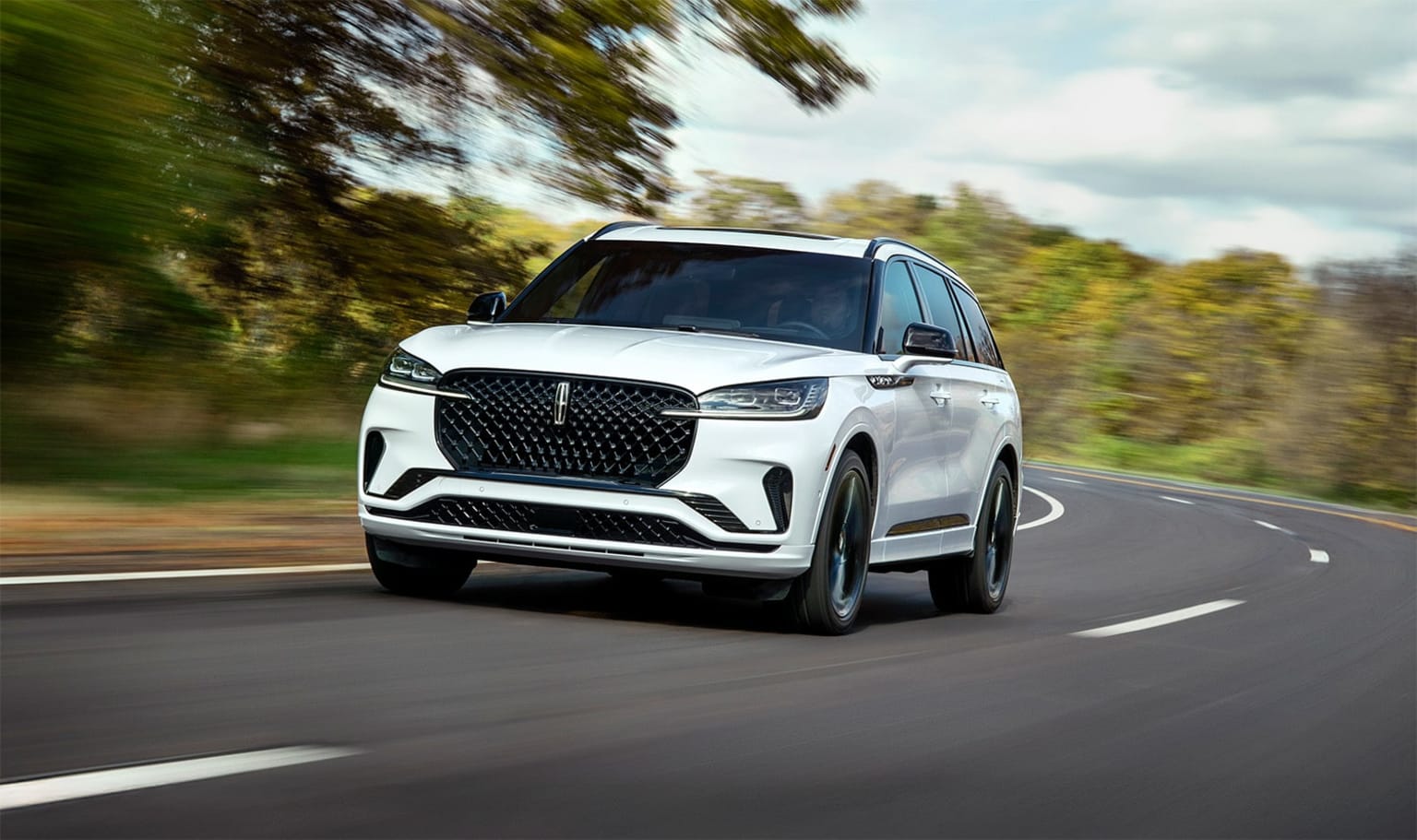A sleek, white SUV driving on a winding road surrounded by lush, blurred foliage in the background.