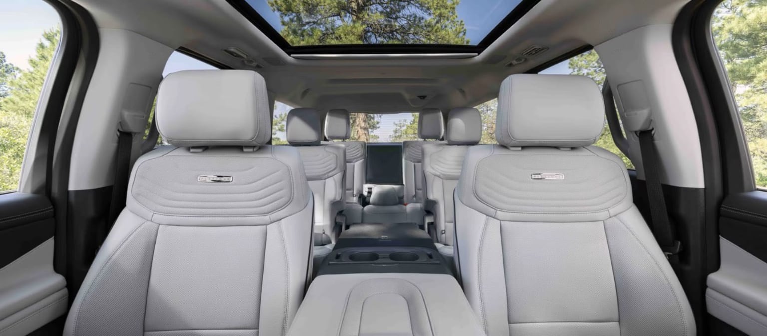 The image shows the interior of a vehicle with a panoramic sunroof visible in the background and rows of comfortable-looking gray seats in the foreground.