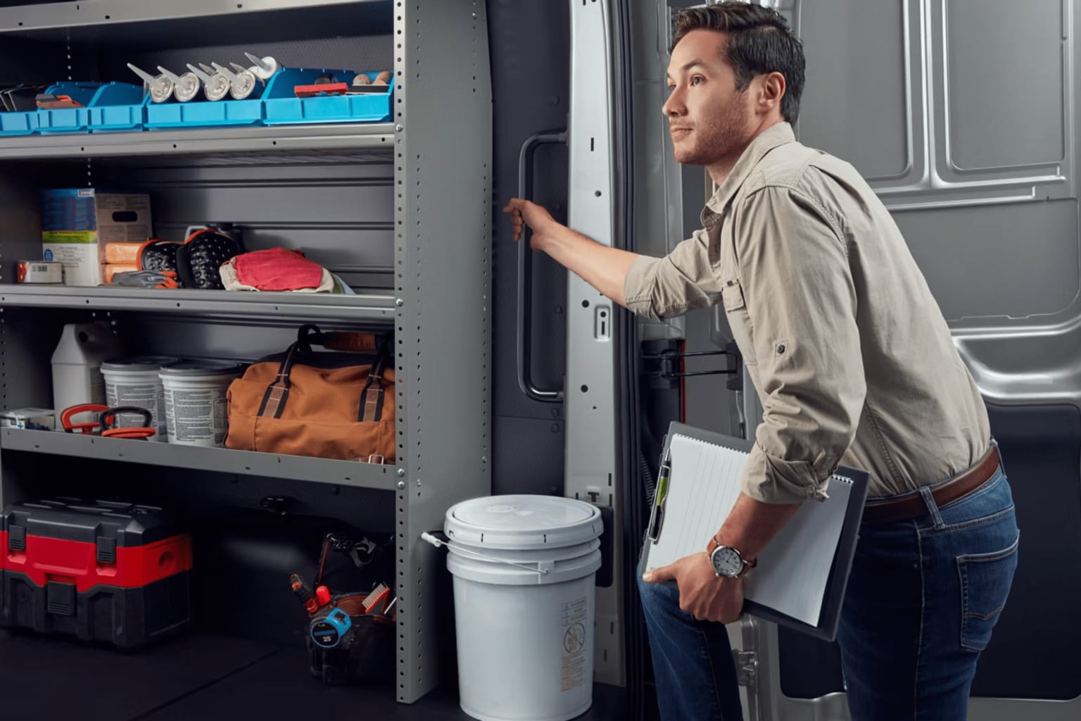 An individual in a tan shirt and jeans, with a clipboard in hand, opening the door of a van stocked with neatly arranged tools and supplies.