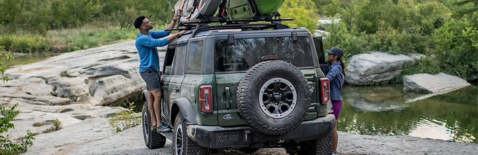 An off-road vehicle with a roof rack and large tires, parked in a natural setting with lush greenery and rocky terrain in the background.