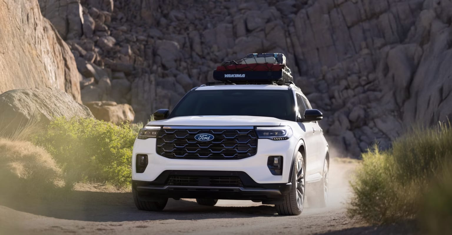 A white SUV with a roof rack drives through a dusty canyon surrounded by rocky cliffs.