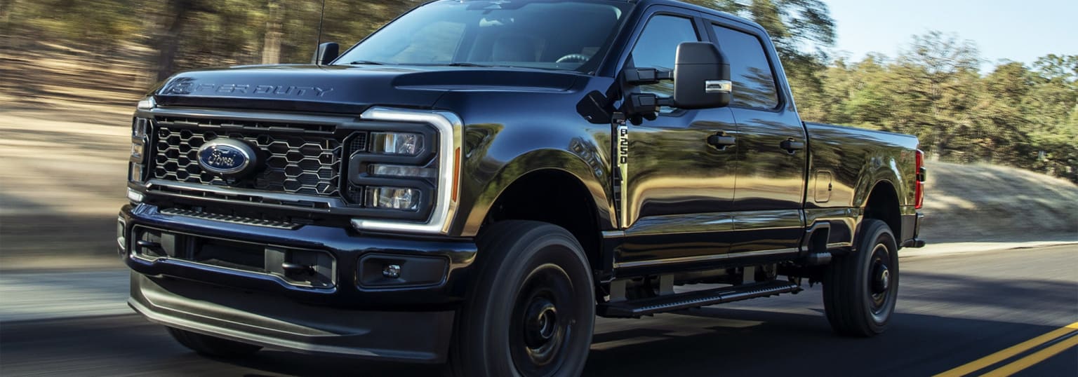 A black, rugged-looking pickup truck with a prominent grille and large tires is speeding through a forested area, with blurred trees and foliage in the background.