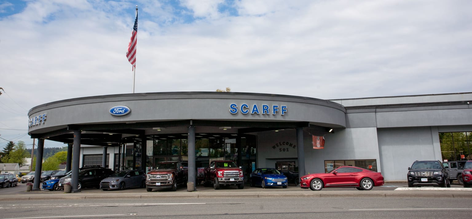 A large gray building with the dealership logo on the front, surrounded by various vehicles including cars and trucks, with an American flag flying atop the structure against a cloudy sky.