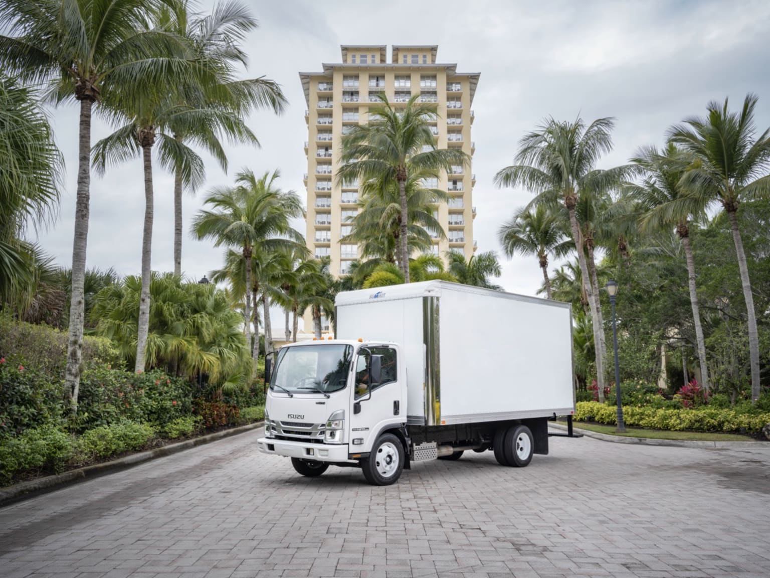 A white delivery truck parked on a paved road with lush palm trees and a tall, multi-story building in the background.