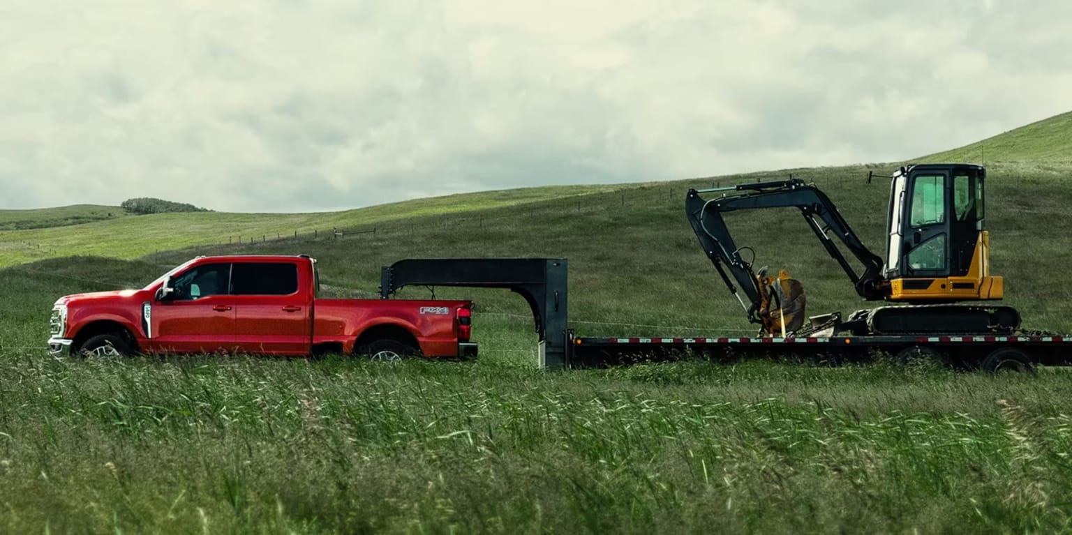 Red pickup truck towing a flatbed trailer with a yellow excavator in a grassy field with rolling green hills in the background
