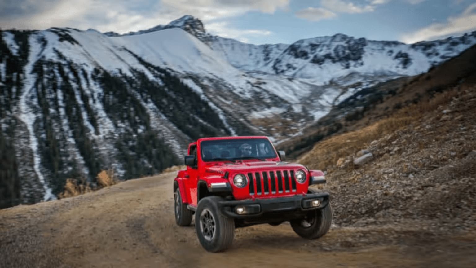 A red SUV traversing a dirt road in a mountainous, snowy landscape with towering peaks in the background.