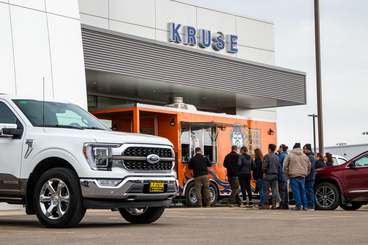 a group of people standing in front of a white truck in front of a kruse store with a red truck parked in front of it