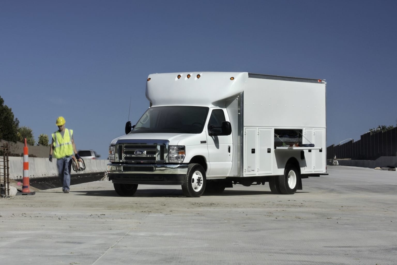 A white utility truck with a large cargo box is parked on a paved road, with a construction worker wearing a safety vest visible in the background.