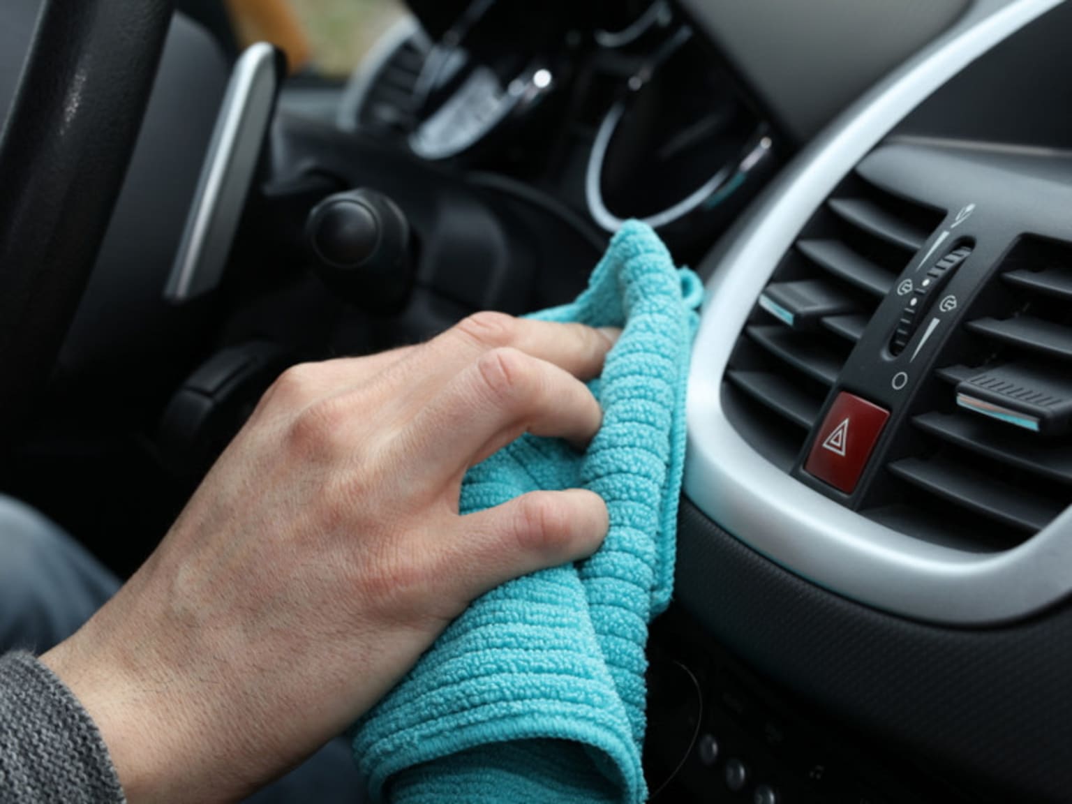 A hand holding a teal-colored microfiber cloth is wiping the dashboard of a car, with various controls and vents visible in the background.
