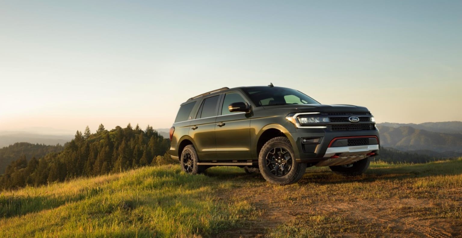 a black suv parked on top of a lush green hillside next to a forest in the distance with a mountain range in the background