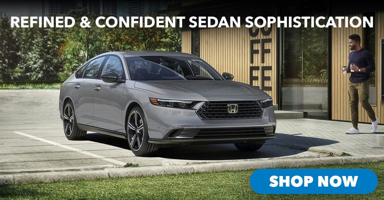 A sleek gray sedan is parked in a lot with a man walking by a coffee shop in the background.