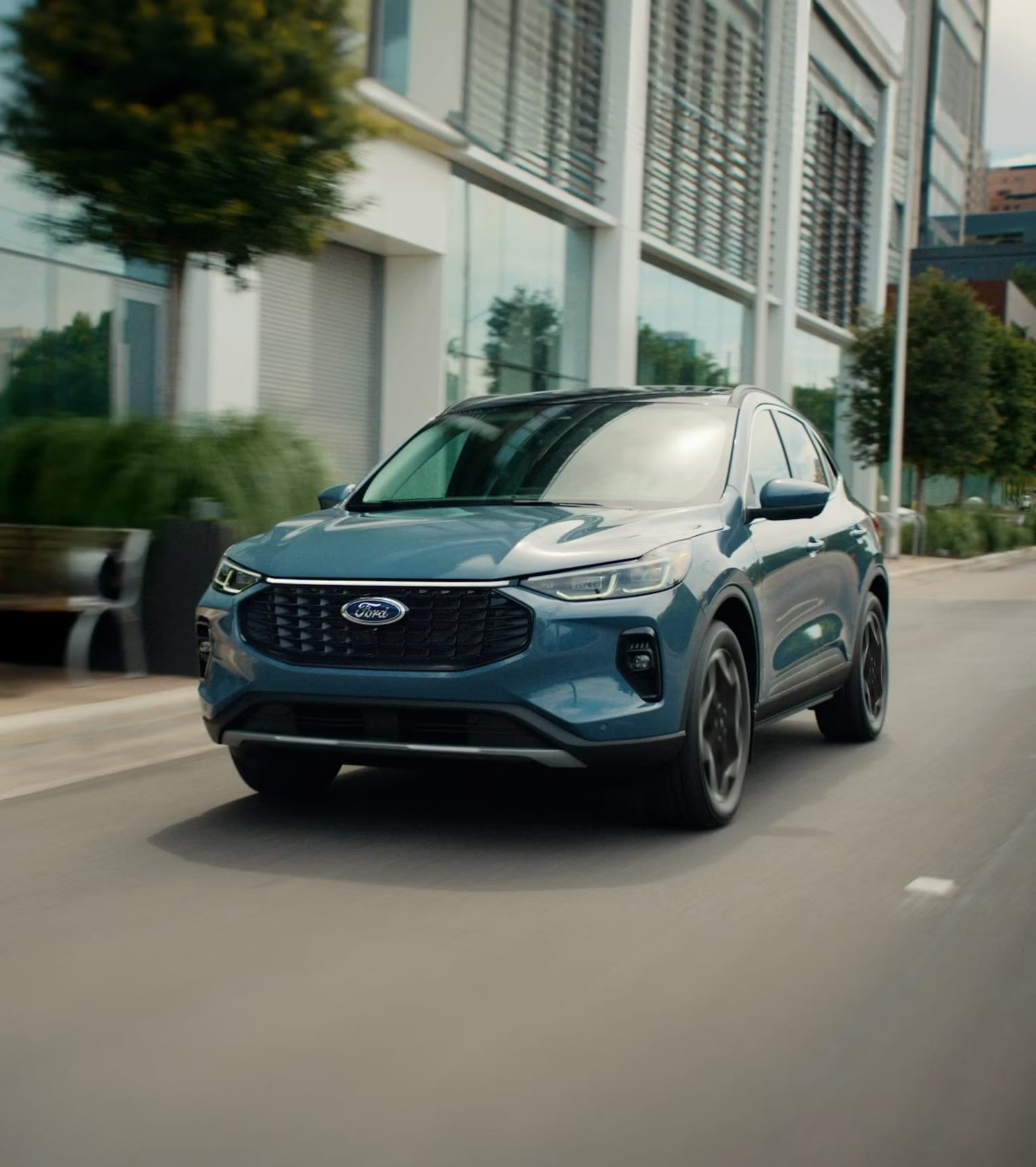 A teal SUV driving down a city street with buildings and greenery in the background.