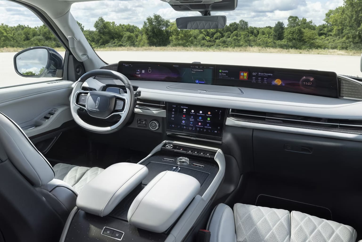 Interior of a modern vehicle with a sleek and technologically advanced dashboard, large display screen, and various controls. Outside view includes trees and greenery visible through the windshield.