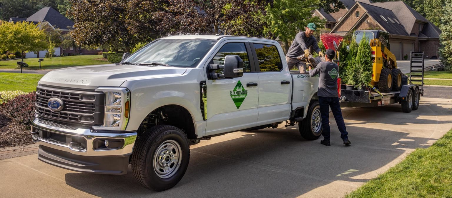 A large white pickup truck with a trailer attached parked on a paved driveway surrounded by lush greenery and residential buildings in the background.