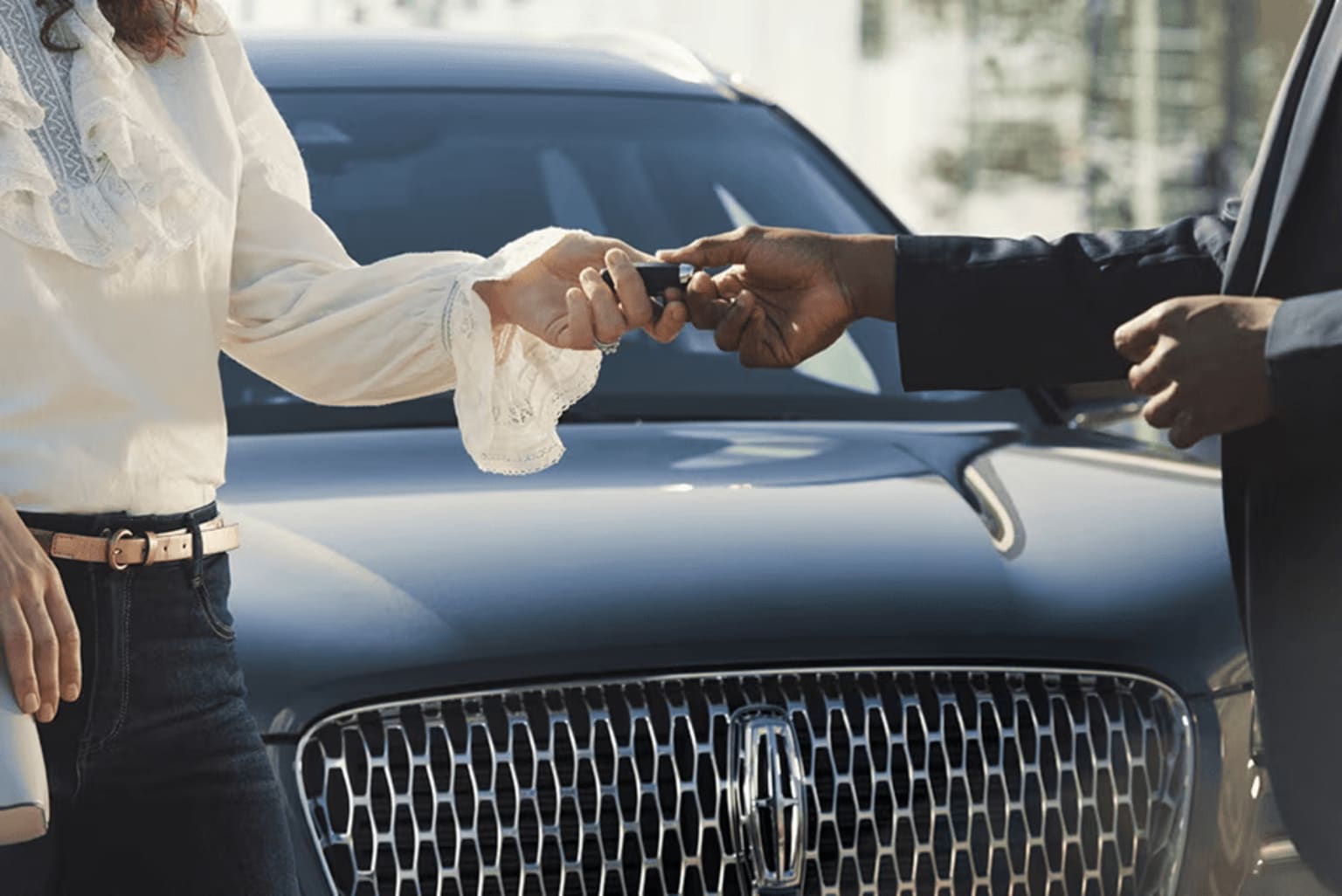 A close-up view of two people's hands shaking in front of a black luxury car in an urban setting.