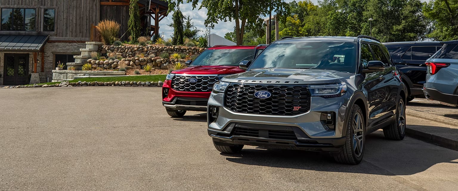 A silver SUV and a red SUV are parked on a paved driveway in front of a rustic wooden house with lush green trees.