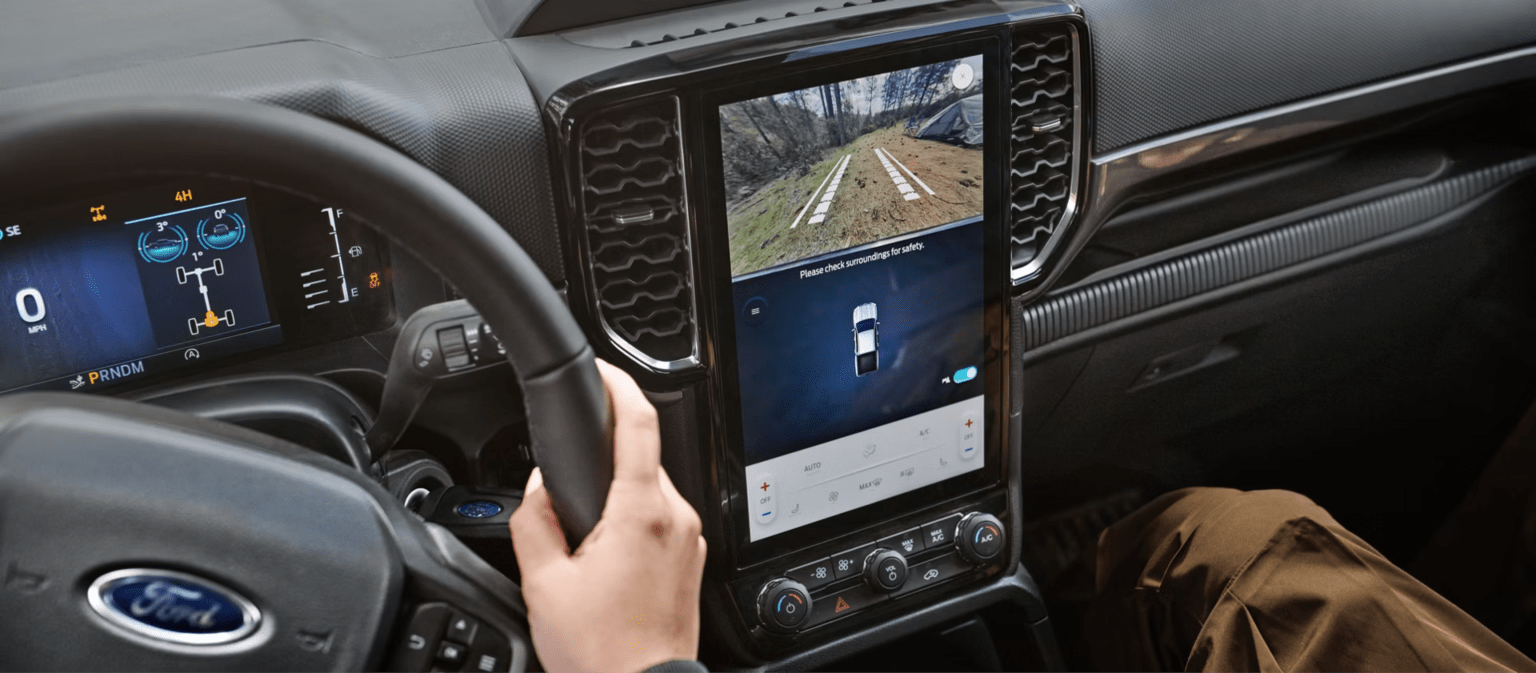 A person's hands grip the steering wheel of a truck, with a large touchscreen displaying a rearview camera feed of a dirt road in the background.