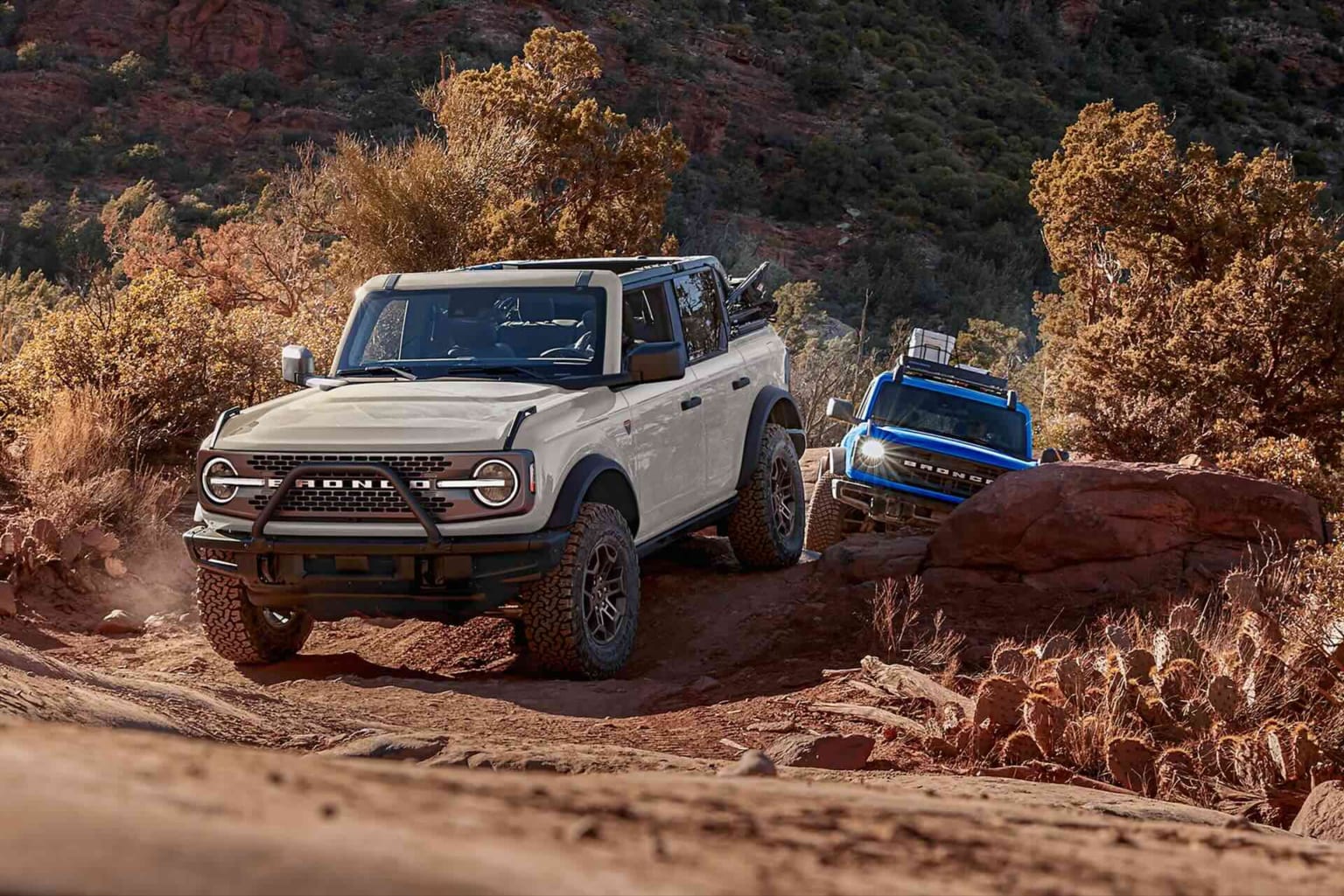 Two SUVs, one white in the foreground and one blue in the background, navigate a rocky, desert terrain with scrubby vegetation and hills.