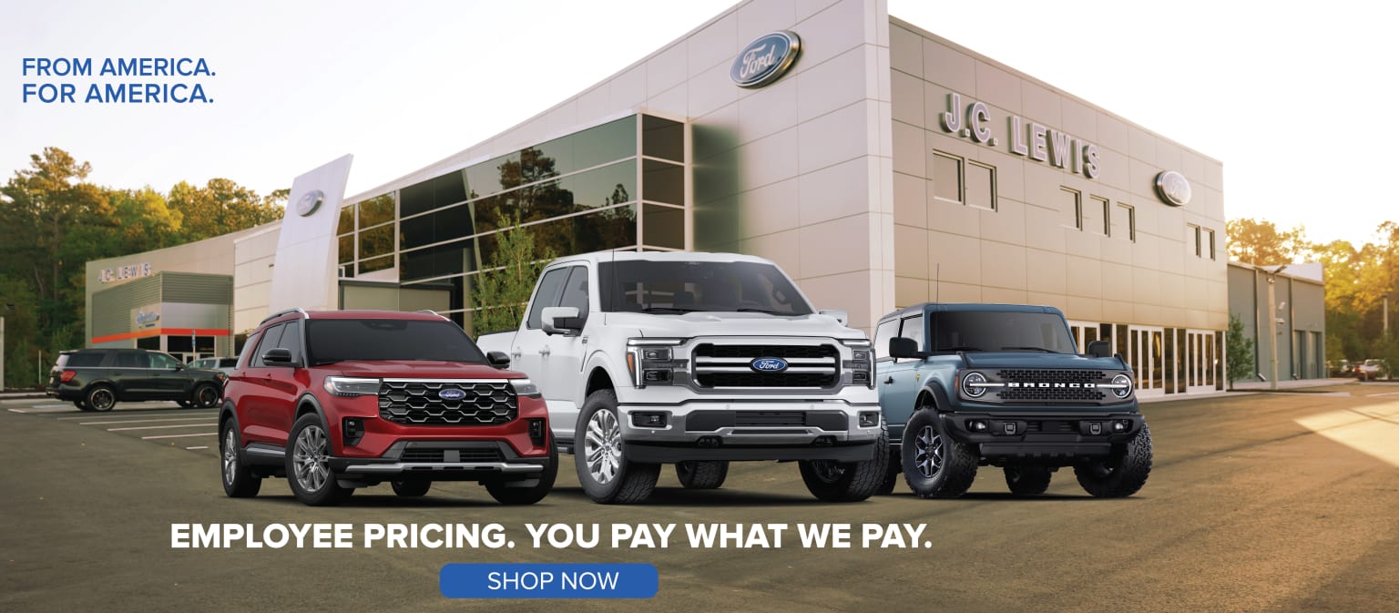 The image depicts a car dealership with various large pickup trucks parked in the foreground, including red, white, and black trucks. The dealership building is shown in the background.