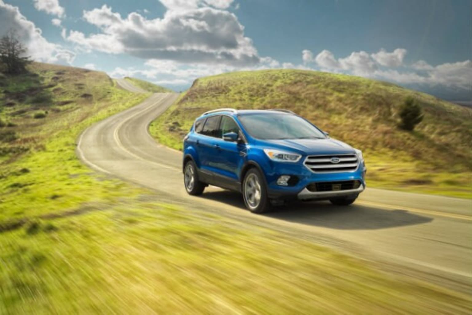 a blue ford escape driving down a country road in the country side with a blue sky and clouds in the background