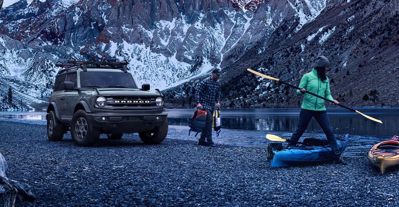 Off-road vehicle parked on a rocky shore with person holding a paddle, snowy mountain landscape in the background.