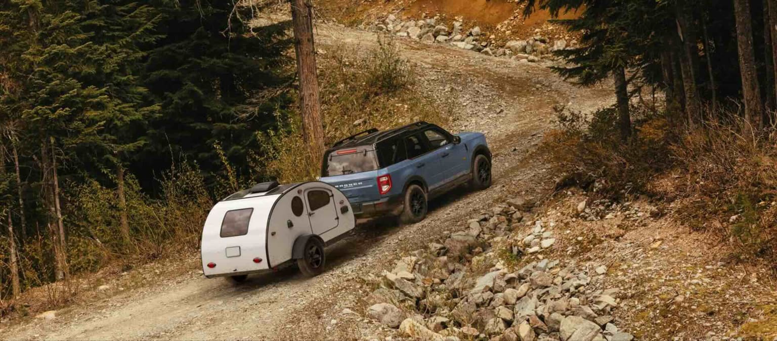 A blue SUV towing a white teardrop trailer up a rocky, wooded mountain road.