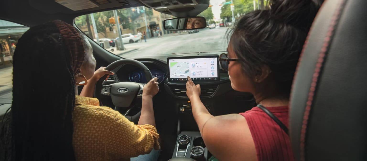 The image shows the interior of a car, with the driver using a navigation app on a tablet-like device mounted on the dashboard. The background outside the car appears to be a city street with buildings and other vehicles visible through the windshield.