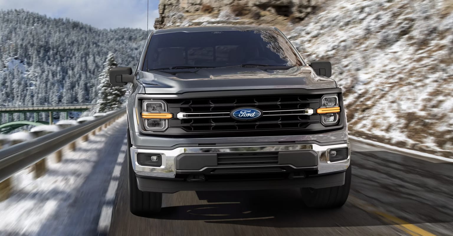 A rugged pickup truck navigates a snowy mountain road, surrounded by towering cliffs and a forested landscape.