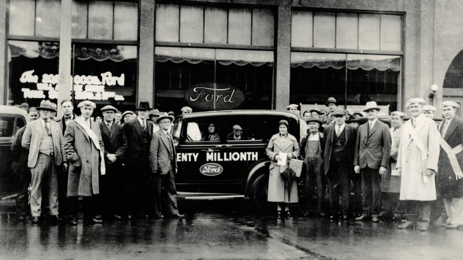 A large group of people, both men and women, standing in front of an automobile with the text "Twenty Millionth" displayed on its side, against the backdrop of a storefront with decorative window displays.