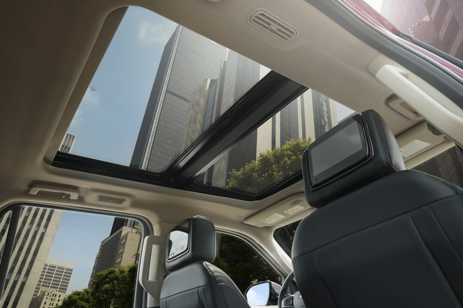 Interior of a vehicle with a large sunroof or panoramic window, offering a view of the surrounding cityscape with tall buildings and greenery in the background.