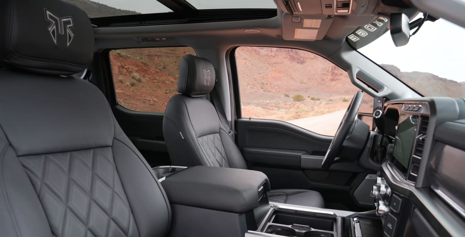 Interior of a vehicle with a dashboard, steering wheel, and seats. Background features a mountainous, rocky landscape.